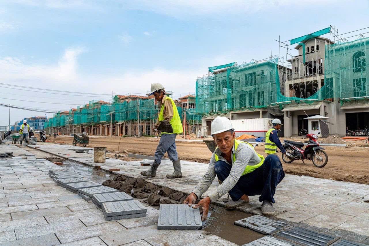 Công nhân đội mũ bảo hộ lát gạch vỉa hè trước dãy nhà đang xây