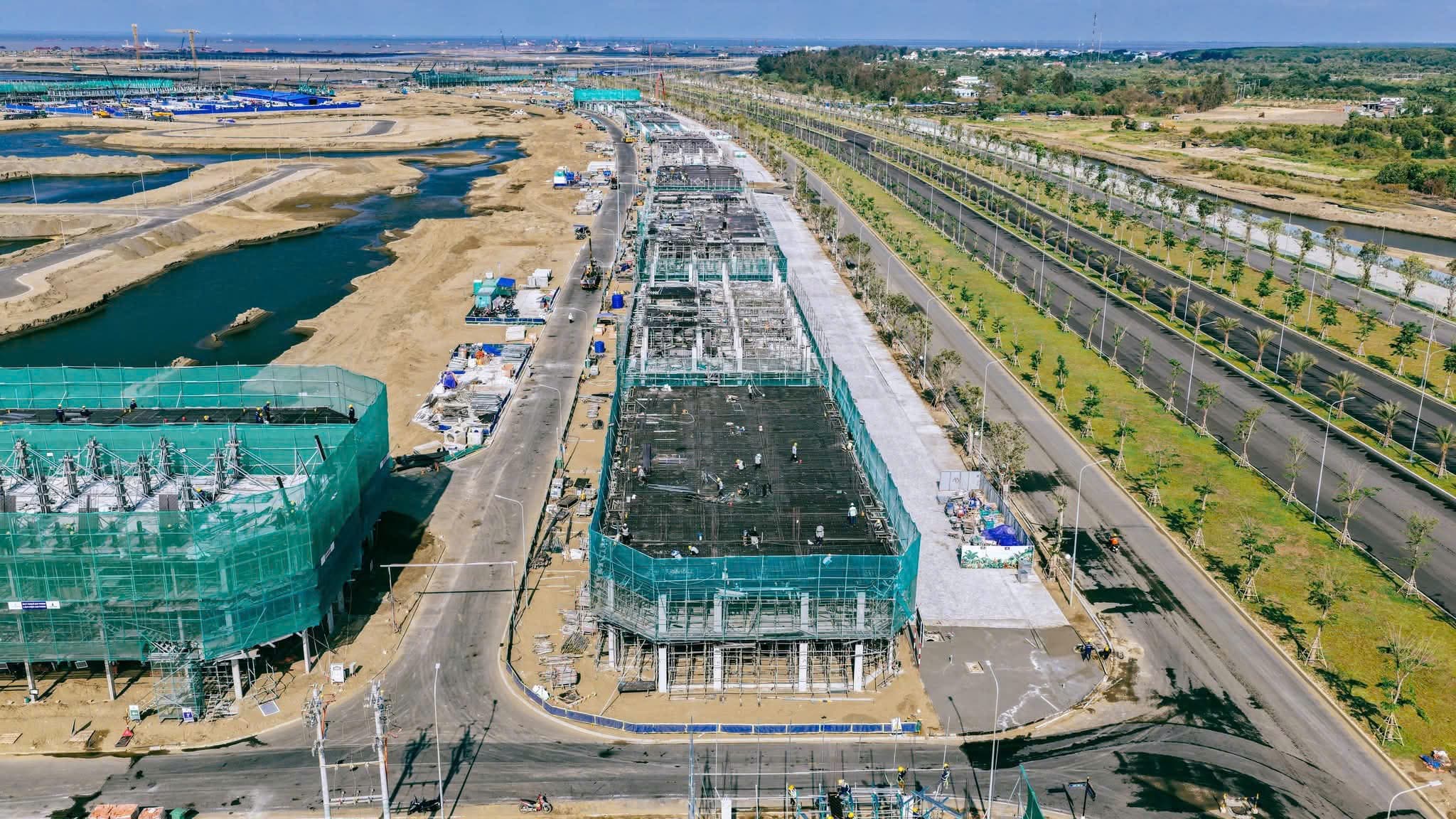 Anh chup tu tren cao day nha pho thuong mai dang xay tho, khung be tong phu luoi gian giao xanh, ben canh la dai lo nhieu lan da trong cay, phia xa la mat nuoc va khu dat dang san lap